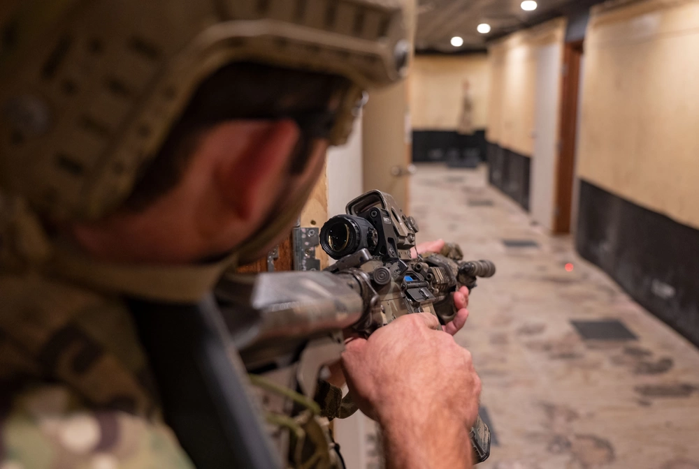 Service member aiming down an indoor training corridor where a faint red laser spot is visible